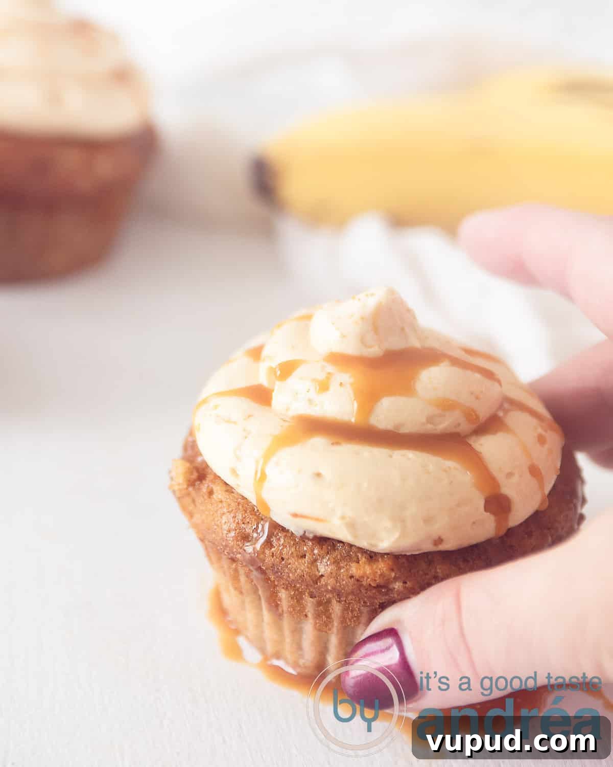 Caramel Banana Cupcakes with Gooey Salted Caramel Drizzle 6 A banana cupcake with caramel cream topping. One hand takes the cupcake. Banana and another cupcake in the background