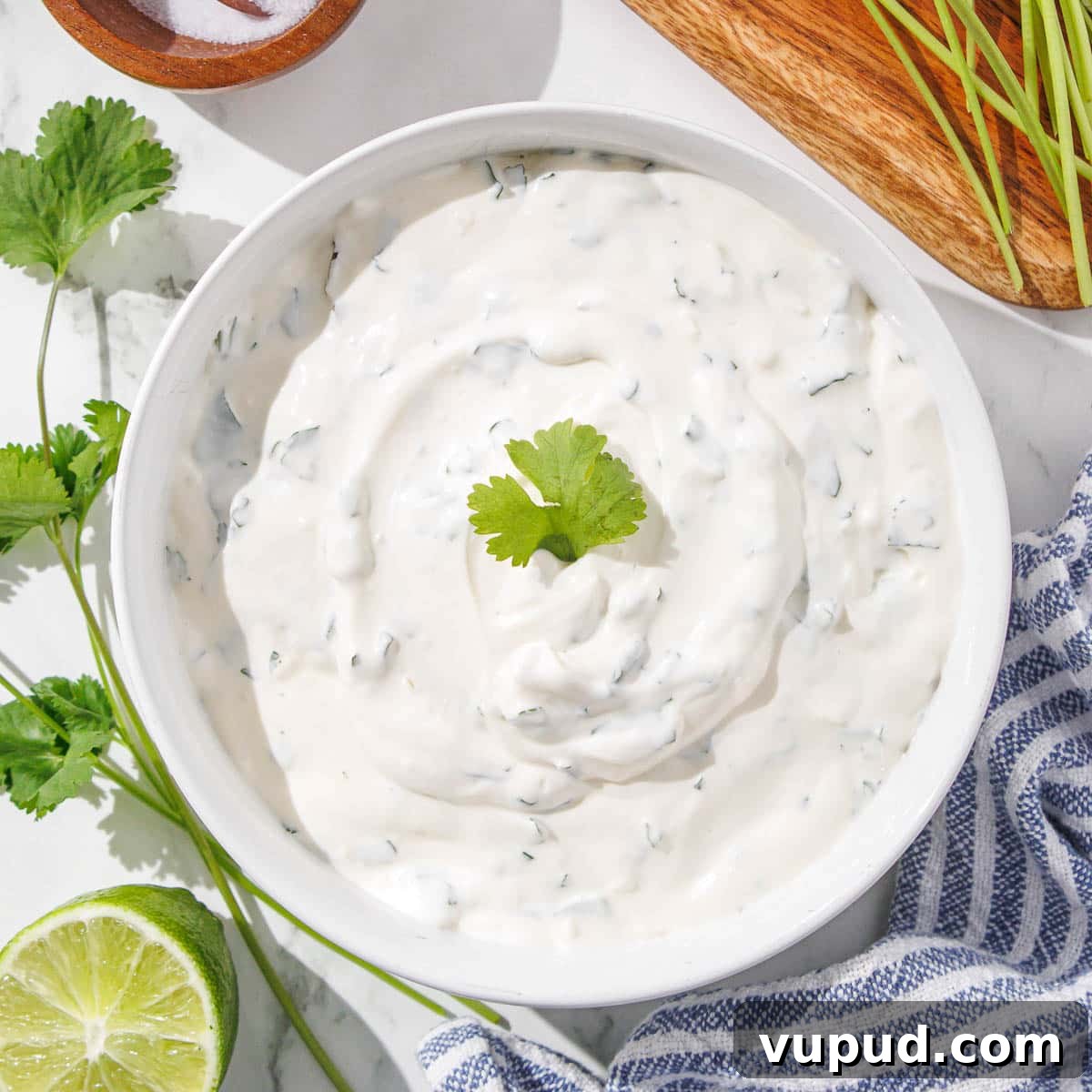 Creamy Cilantro-Lime Crema Recipe for Tacos & Bowls 8 A bowl of homemade cilantro lime crema garnished with a cilantro leaf, surrounded by fresh limes and a wooden spoon.
