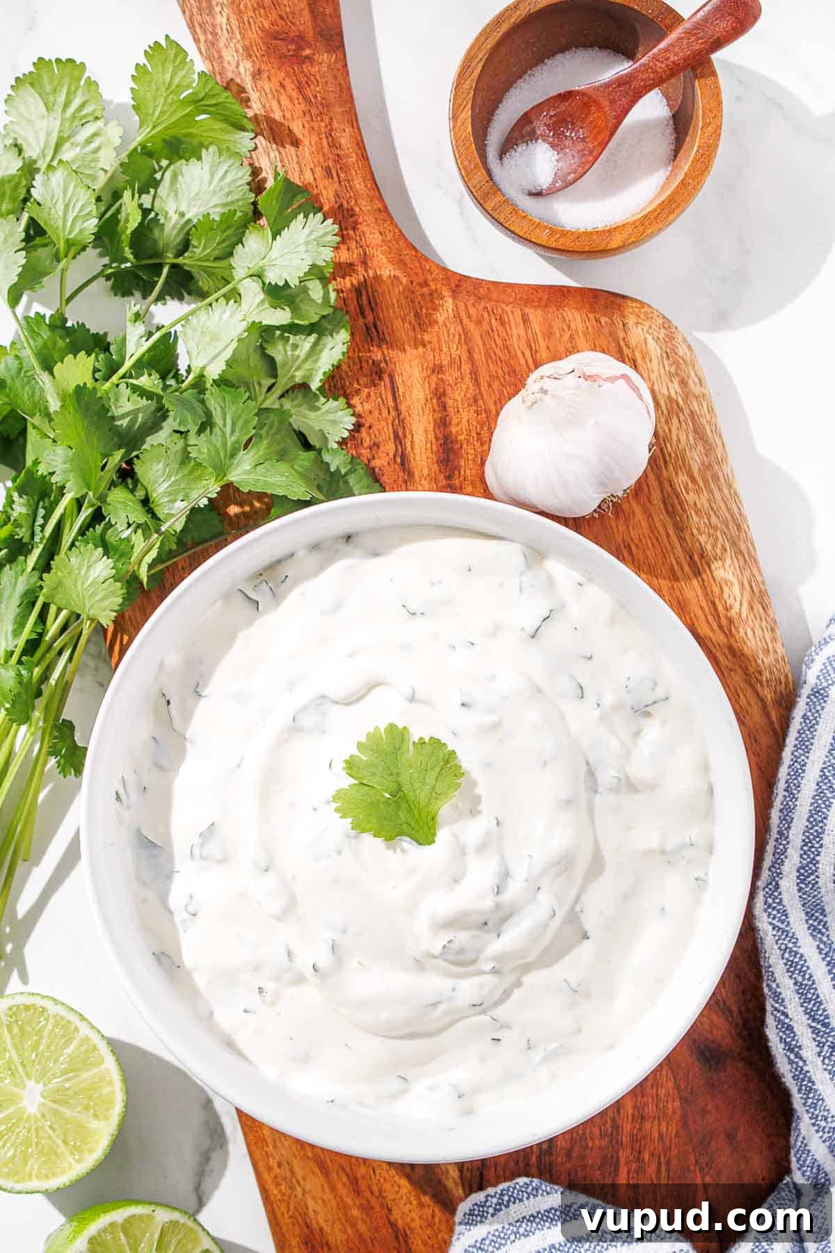 Creamy Cilantro-Lime Crema Recipe for Tacos & Bowls 10 A bowl of cilantro lime crema on a wooden board, flanked by fresh cilantro, lime, garlic, and a pinch bowl of salt.