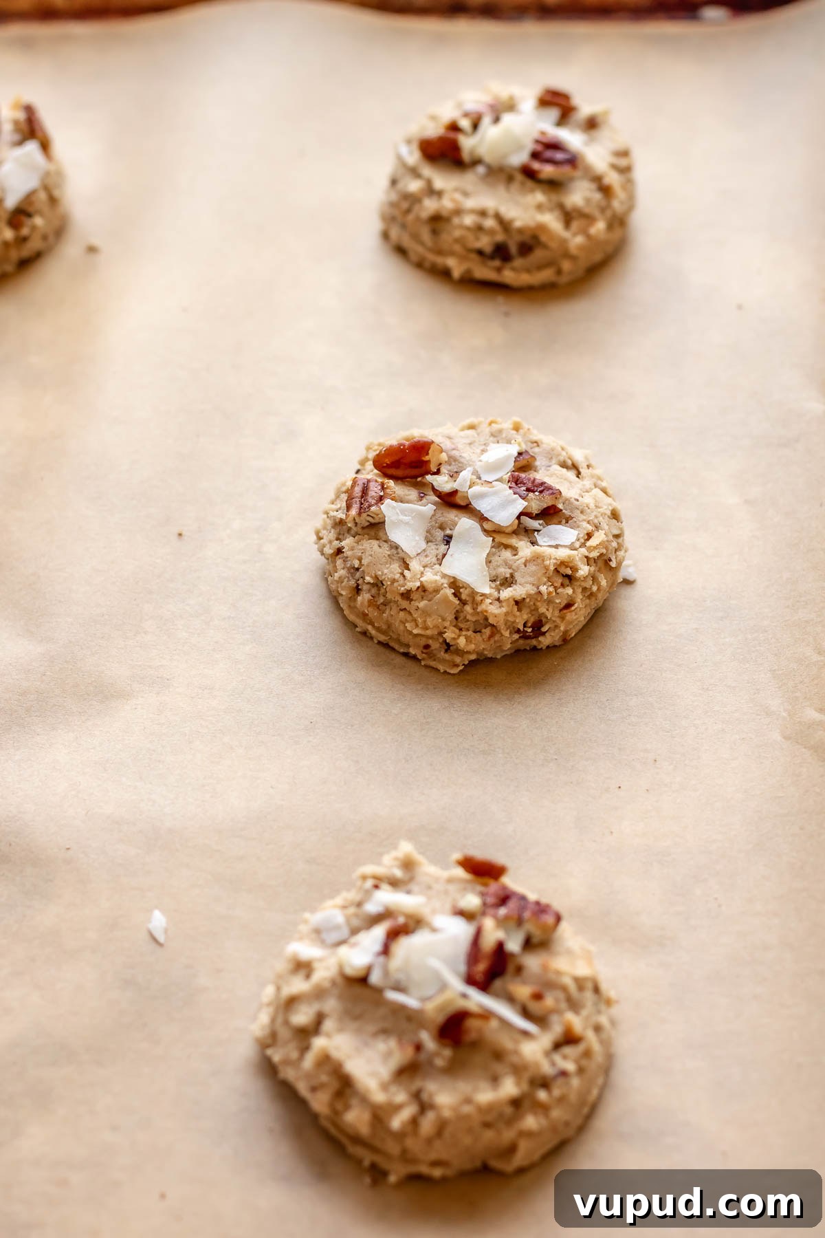 Chewy Coconut Pecan Cookies Recipe for Crispy Edges and Gooey Center 12 Cookie dough on a baking sheet with coconut flakes and pecans on top.