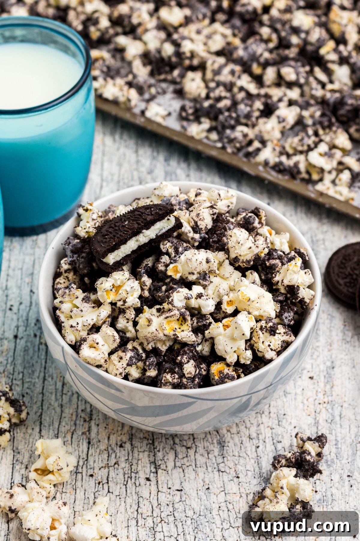 Oreo Popcorn Recipe: Creamy Chocolate Cookie Popcorn Treat 2 Popcorn in a light bowl with crushed Oreo cookies mixed in with white chocolate in front of blue glasses filled with milk.