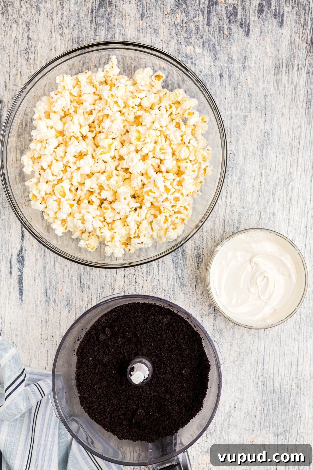 Oreo Popcorn Recipe: Creamy Chocolate Cookie Popcorn Treat 4 Oreos in a food processor bowl, crushed into crumbs, with melted white chocolate and a bowl a popcorn, on a blue wooden table.