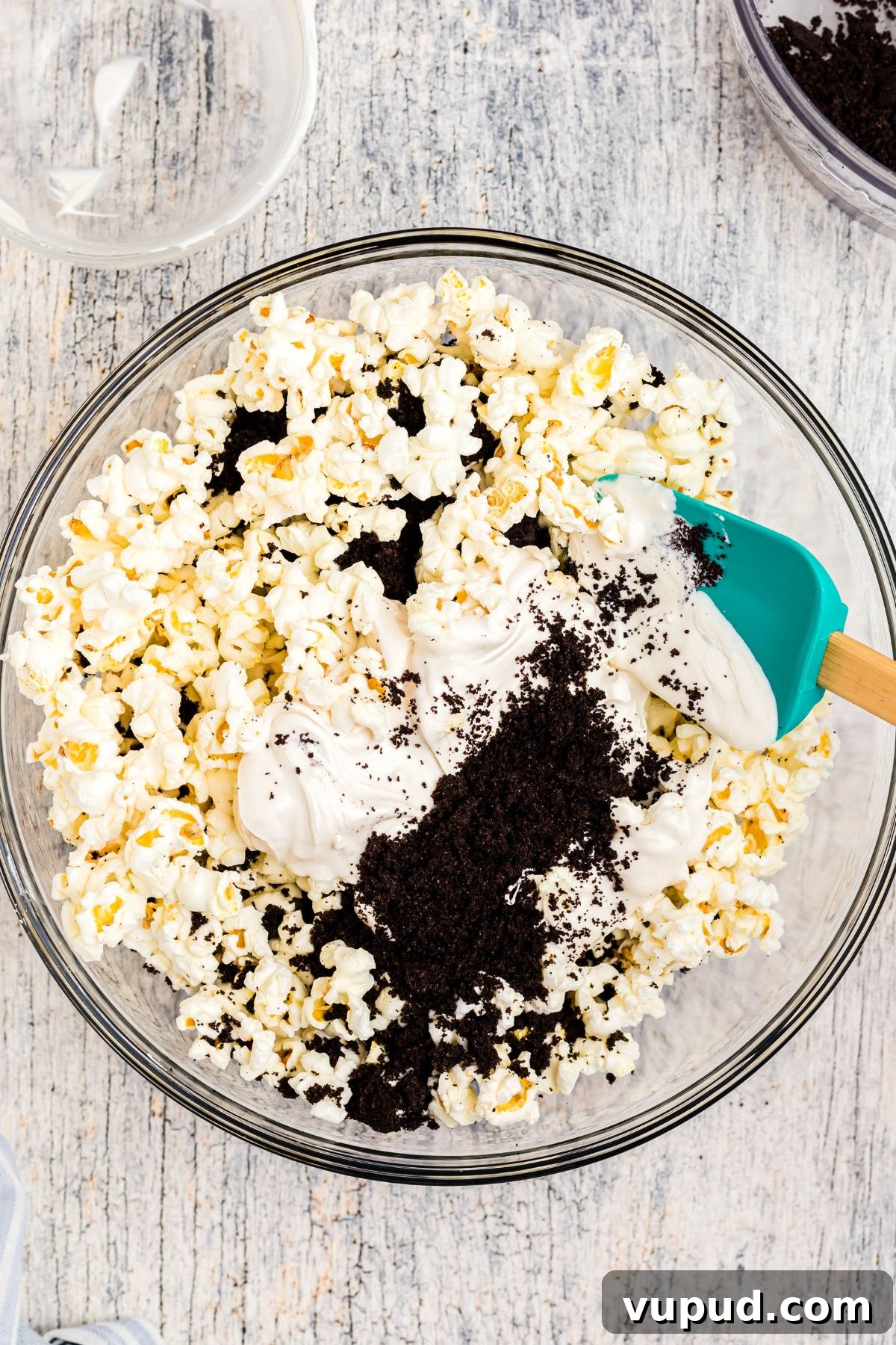 Oreo Popcorn Recipe: Creamy Chocolate Cookie Popcorn Treat 5 Melted white chocolate with Oreo crumbles being mixed in a clear glass bowl filled with popcorn, with a spatula.