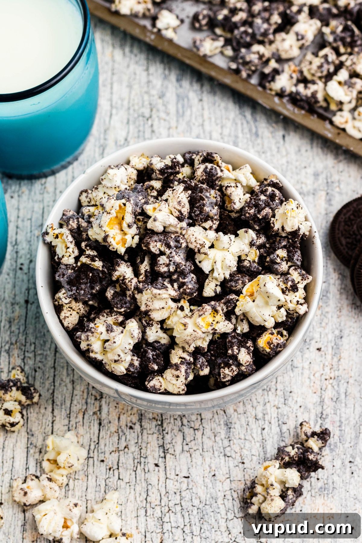 Oreo Popcorn Recipe: Creamy Chocolate Cookie Popcorn Treat 7 A bowl filled with Oreo popcorn on a table with blue glasses filled with milk and popcorn pieces scattered on the table.