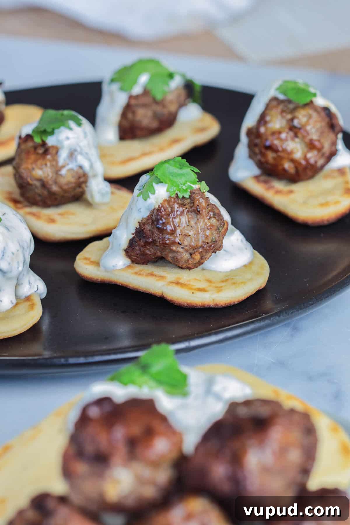 Greek Lamb Meatballs with Tzatziki Sauce Recipe 9 Lamb meatballs over pita bread on black plate with sauce.