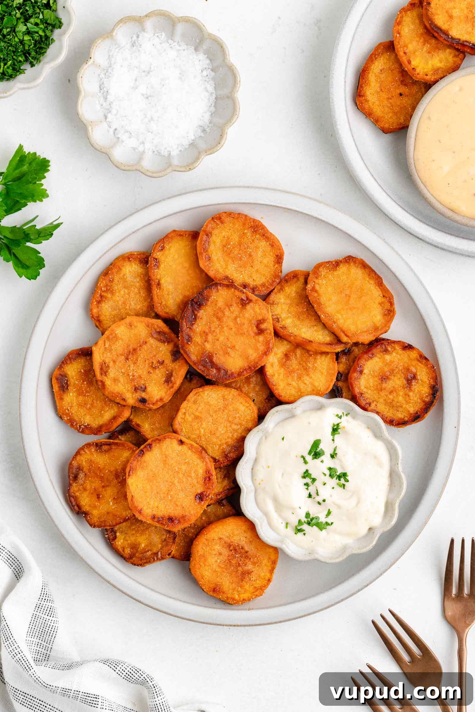 Crispy Fried Sweet Potatoes with Honey-Citrus Glaze 2 A plate of crispy fried sweet potatoes served with a small bowl of creamy dipping sauce, garnished with chopped herbs. Extra sauce, salt, and utensils are nearby on a white table.