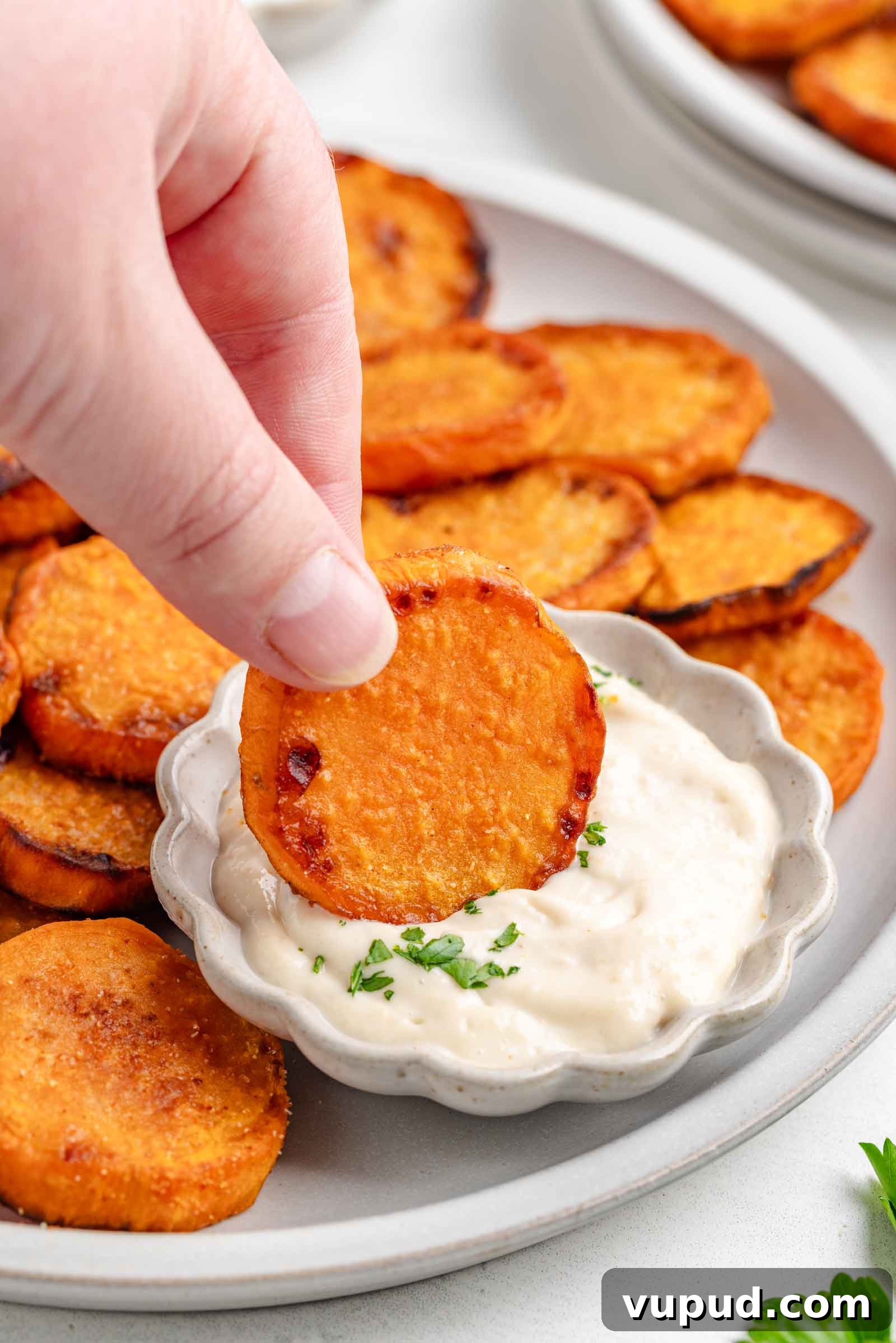 Crispy Fried Sweet Potatoes with Honey-Citrus Glaze 6 A hand dips a round, roasted sweet potato slice into a small bowl of creamy white sauce, with more sweet potato slices arranged on a plate in the background.
