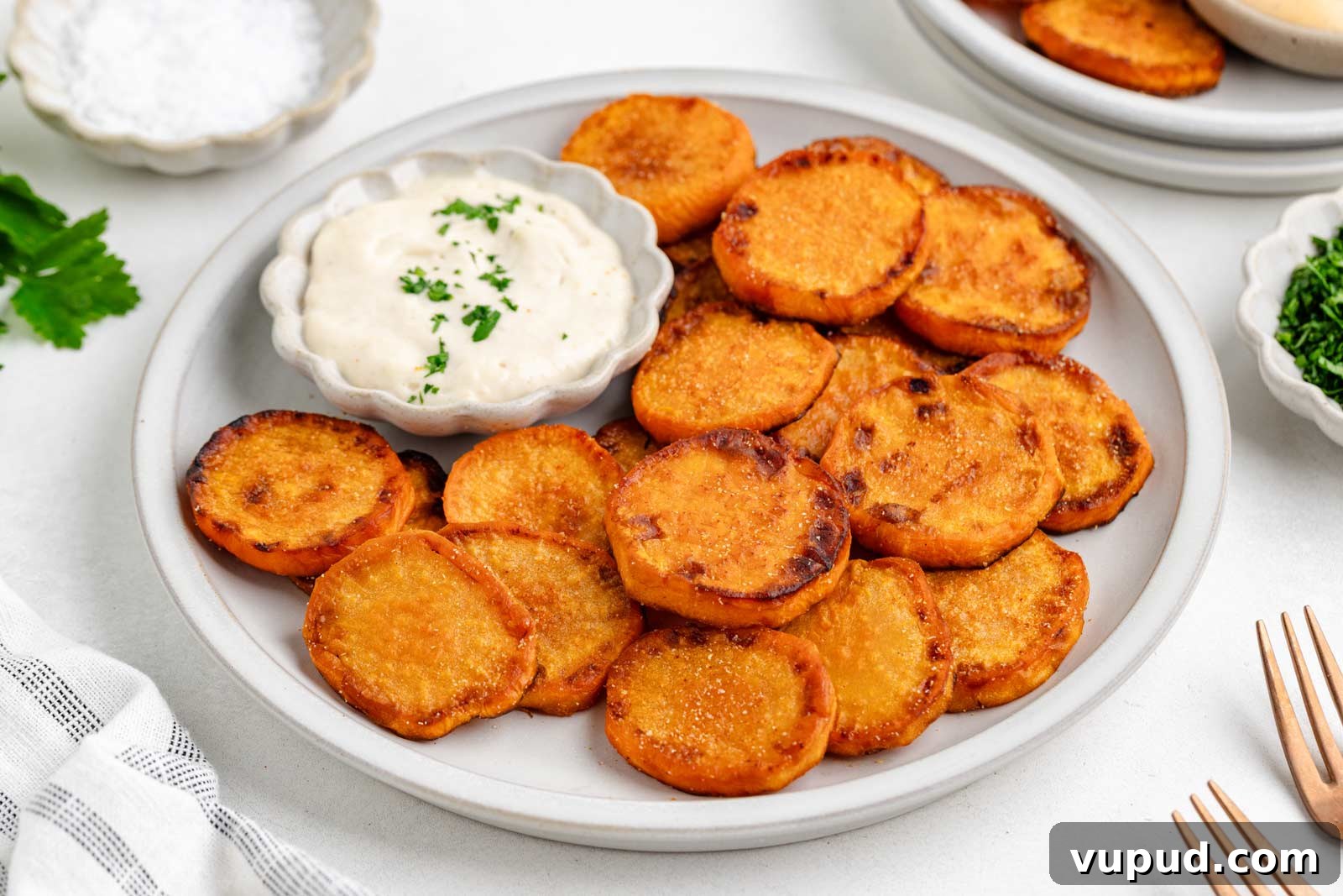 Crispy Fried Sweet Potatoes with Honey-Citrus Glaze 7 A plate of fried sweet potatoes is served with a small bowl of creamy dipping sauce topped with chopped herbs. A fork, napkin, and small bowls of salt and herbs are nearby.