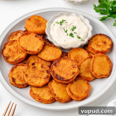 Crispy Fried Sweet Potatoes with Honey-Citrus Glaze 8 A plate of fried sweet potatoes slices served with a small dish of creamy white dipping sauce, garnished with chopped herbs. Fresh parsley and a fork are visible nearby.