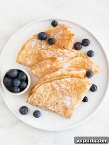 Overview of a plate with several folded vegan crepes topped with fresh blueberries and powdered sugar.
