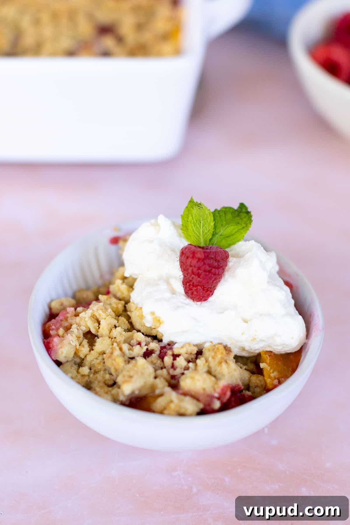 How to Make Perfect Homemade Whipped Cream at Home 3 A bowl of fruit crisp topped with homemade whipped cream, garnished with a raspberry and a mint leaf, placed on a light-colored surface.