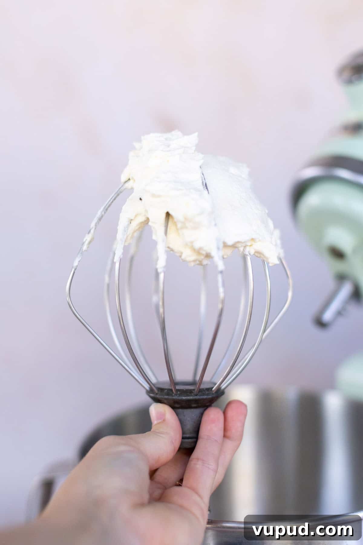 How to Make Perfect Homemade Whipped Cream at Home 7 A hand holding a stand mixer whisk attachment with homemade whipped cream clinging to it. A stand mixer is visible in the background.