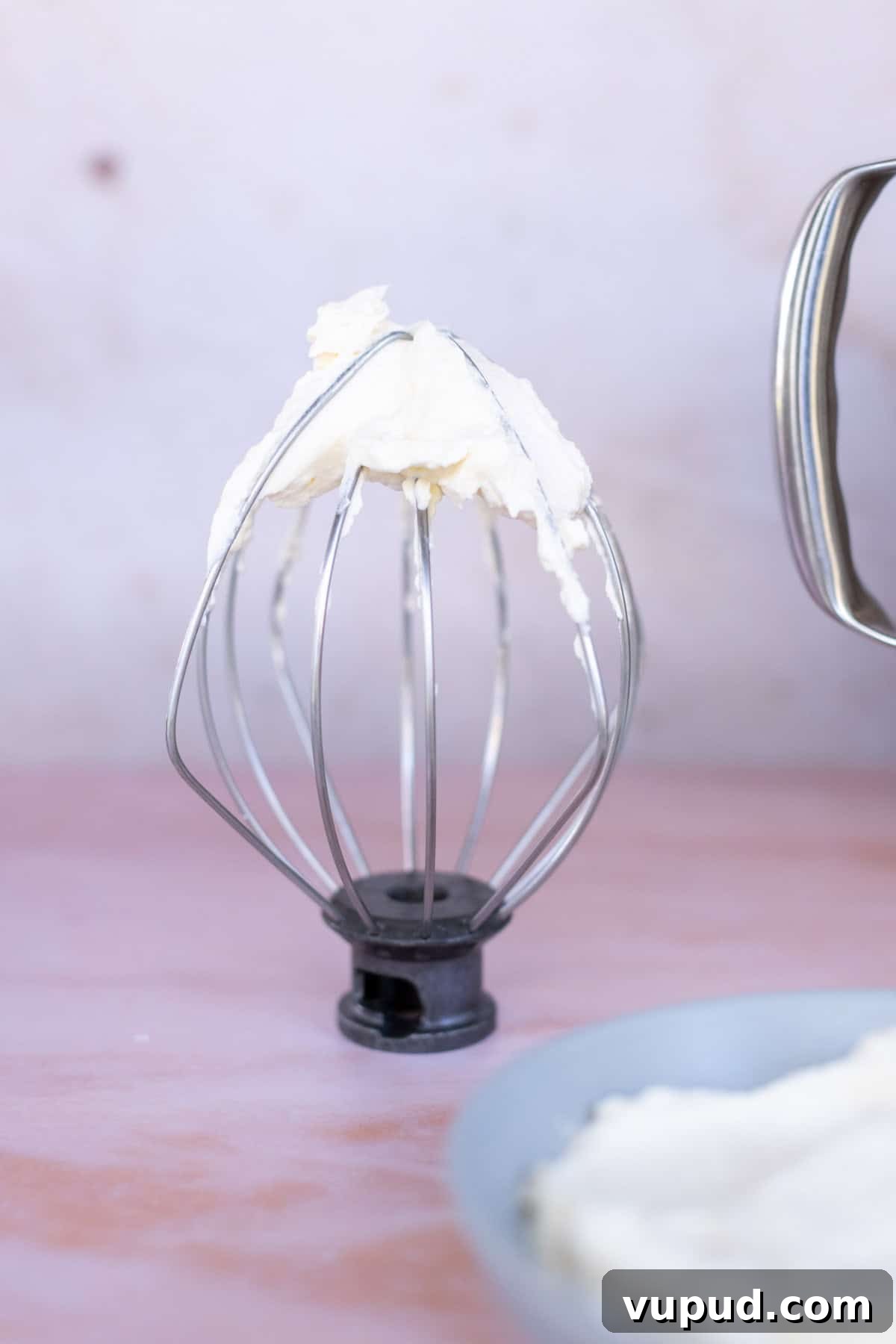 How to Make Perfect Homemade Whipped Cream at Home 9 Close-up of a mixer whisk attachment with homemade whipped cream adhered to its wires, placed on a light pink surface. A partially visible bowl with more whipped cream is in the foreground.