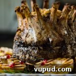 A cooked crown roast of pork sits on a white serving tray with grilled pears and fresh rosemary around the roast.