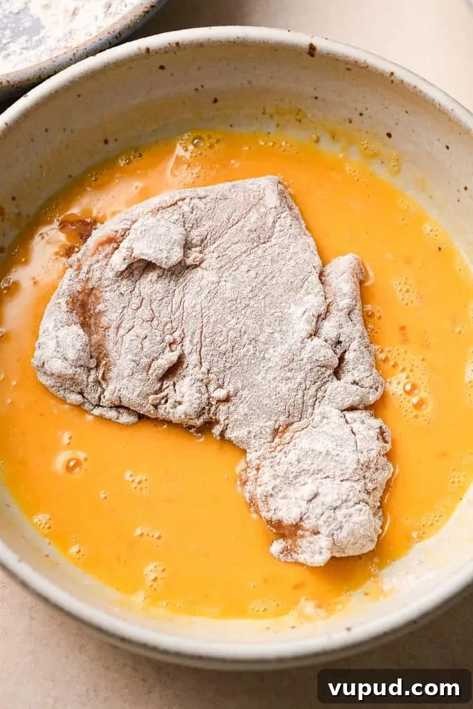 chicken schnitzel recipe A piece of chicken dredged in flour, being dipped into a bowl of beaten eggs.