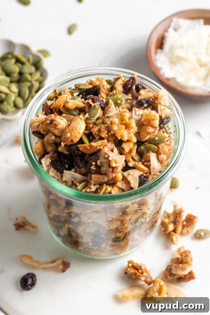 Grain-Free Homemade Granola Recipe for Crunchy, Low-Carb Mornings 4 nut and seed grain free granola in a clear french mason jar with two smaller bowls of pepitas and unsweetened coconut flakes on the white table and some of the granola and seeds scattered around.