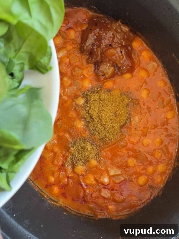 Slow-Cooked Chickpea and Spinach Curry (Chana Saag) 6 A slow cooker pan filled with chickpeas in a sauce, with spices on top and spinach about to the tipped in.