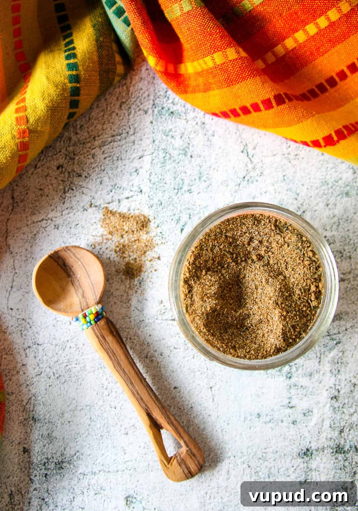 A small wooden spoon next to a glass jar full of Jamaican jerk seasoning blend.