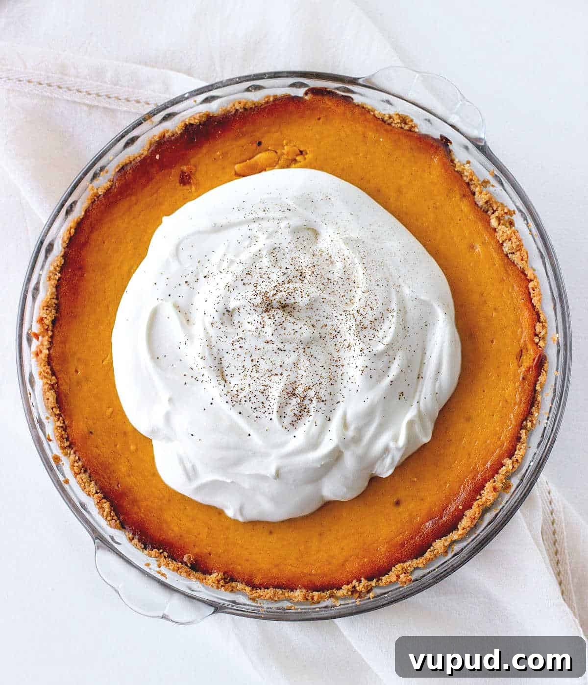 Pumpkin Pie with Graham Cracker Crust Recipe for Fall Baking 3 Top view of whole pumpkin pie with cream on top on a glass dish. White cloth and surface.