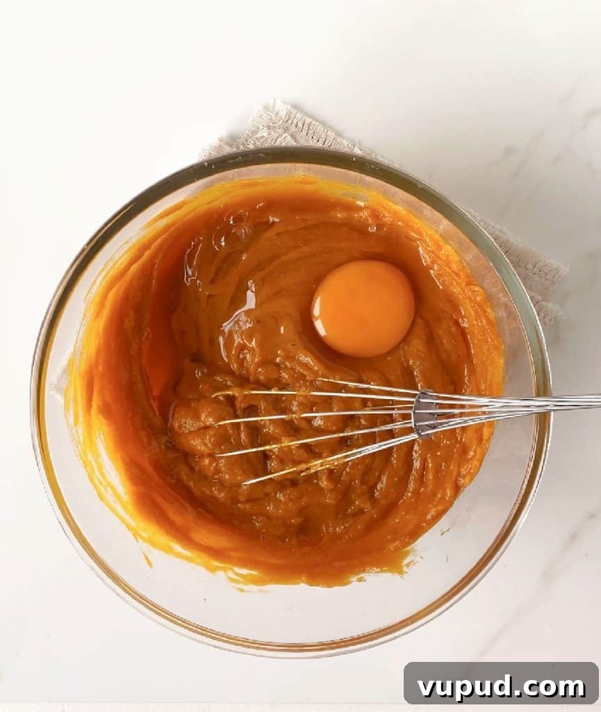 Pumpkin Pie with Graham Cracker Crust Recipe for Fall Baking 8 Egg added to pumpkin pie filling in a glass bowl with a whisk. White marble surface.