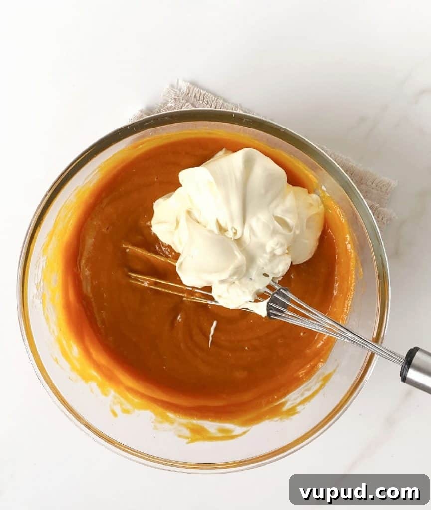 Pumpkin Pie with Graham Cracker Crust Recipe for Fall Baking 9 Sour cream and cream added to pumpkin pie filling in a glass bowl with a wire whisk. White marble surface.