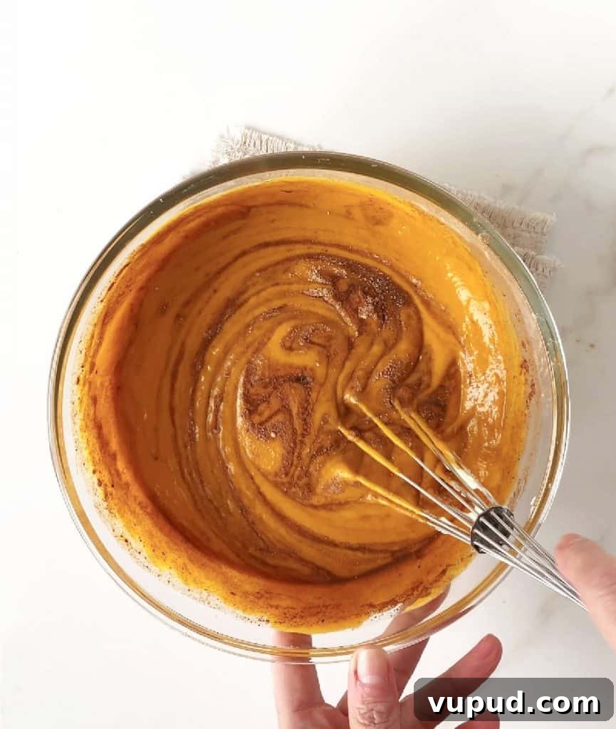 Pumpkin Pie with Graham Cracker Crust Recipe for Fall Baking 10 Top view of pumpkin pie filling with spices being whisked in a glass bowl on a white surface.