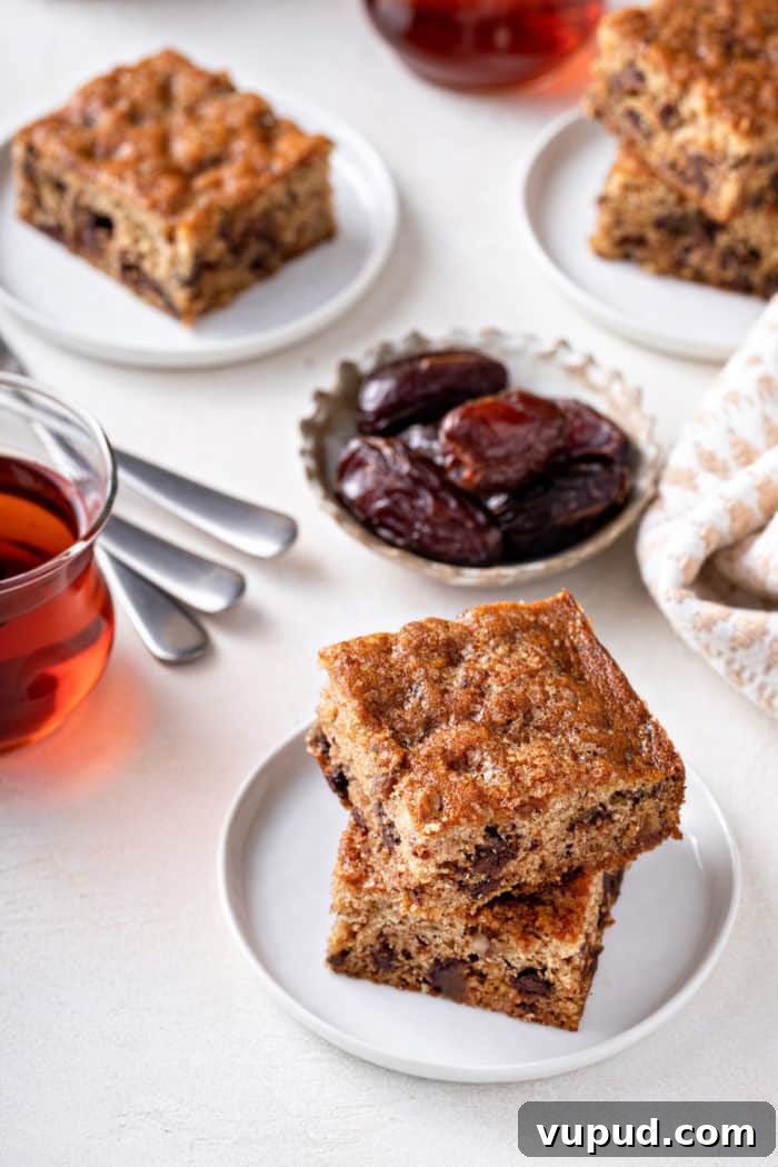 Decadent Date Cake Recipe with Warm Spices 8 Two slices of date cake stacked on a white plate with more plates of cake visible in the background.