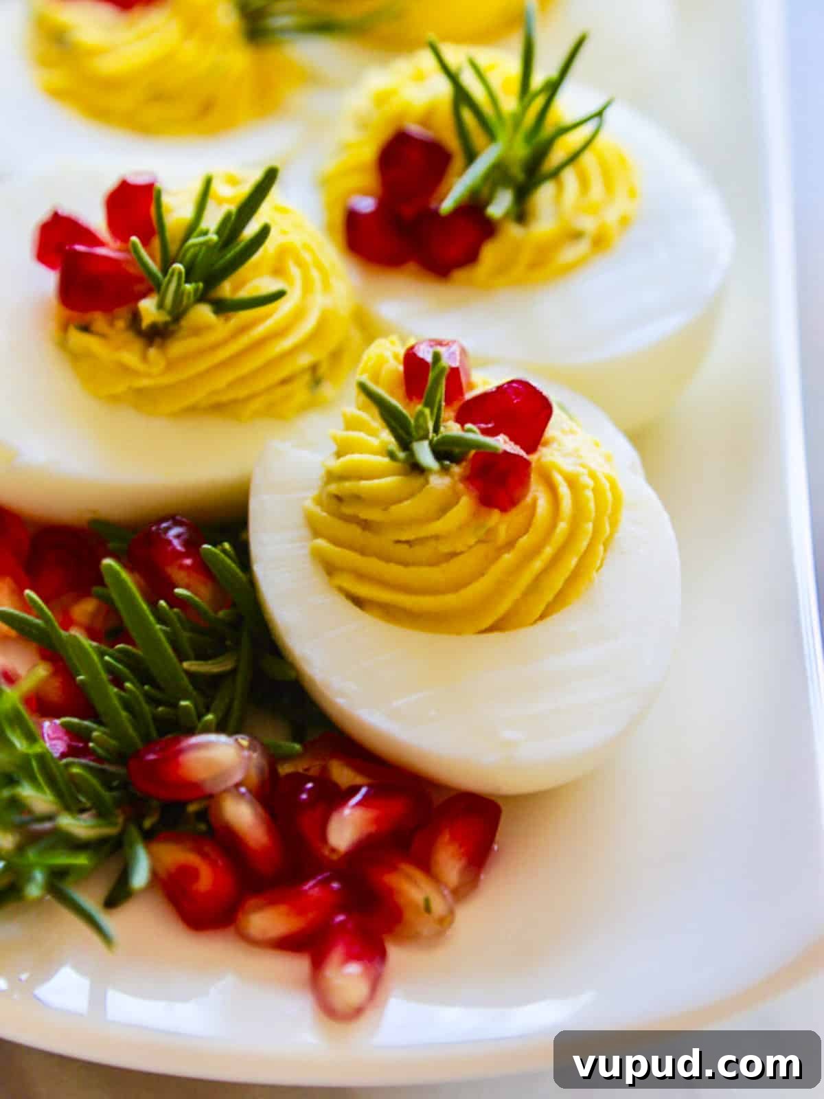 Festive Christmas Deviled Eggs Recipe for Holiday Parties 10 White long platter with gorgeous Christmas Deviled Eggs decorated with rosemary and pomegranate seeds.