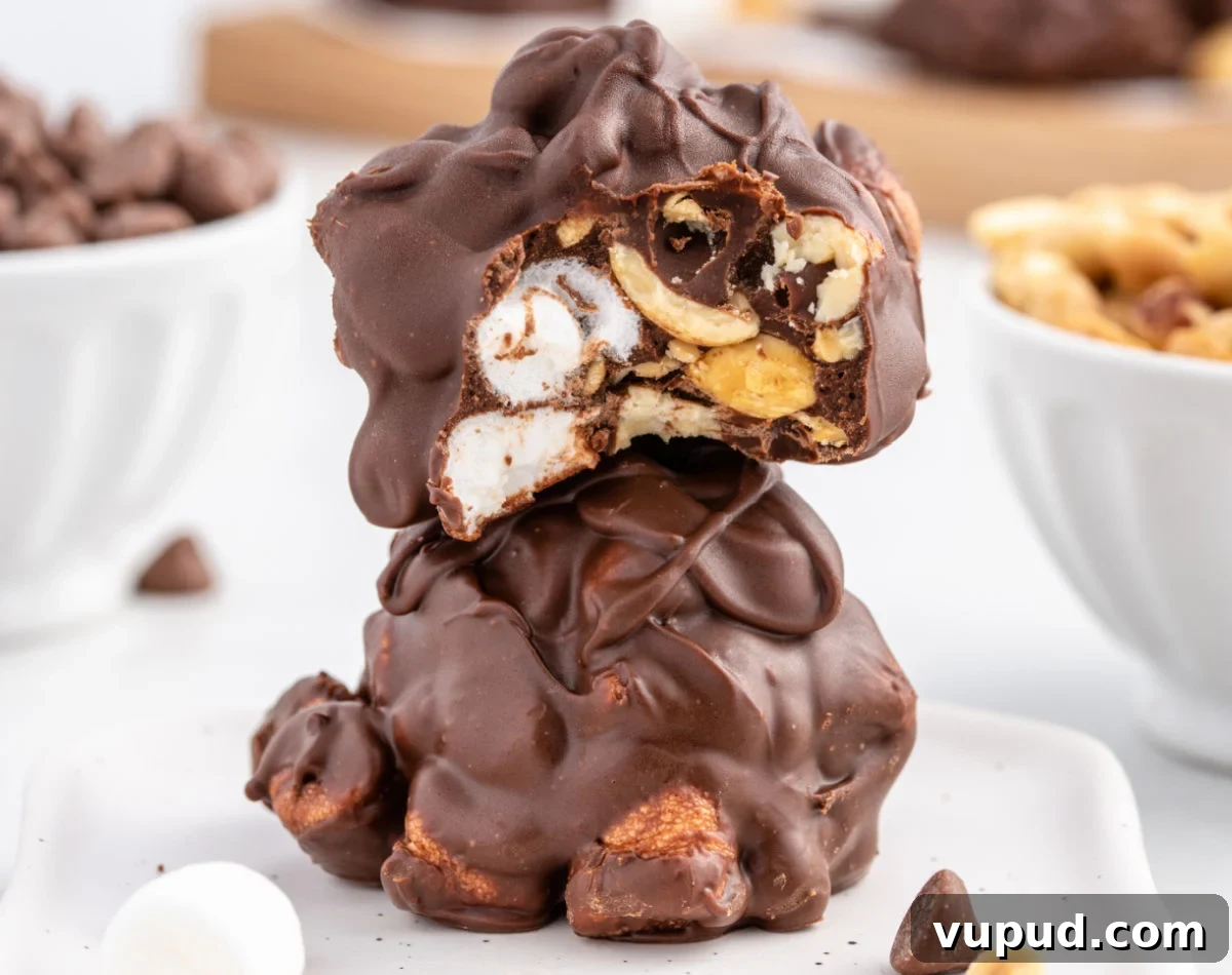 Peanut and Marshmallow Cluster Recipe: No-Bake Candy Clusters 7 A close-up of two stacked peanut marshmallow clusters coated in glossy chocolate. The top cluster is broken open to reveal whole mini marshmallows, crunchy peanuts, and thick chocolate layers inside, with bowls of chocolate chips and peanuts blurred in the background.