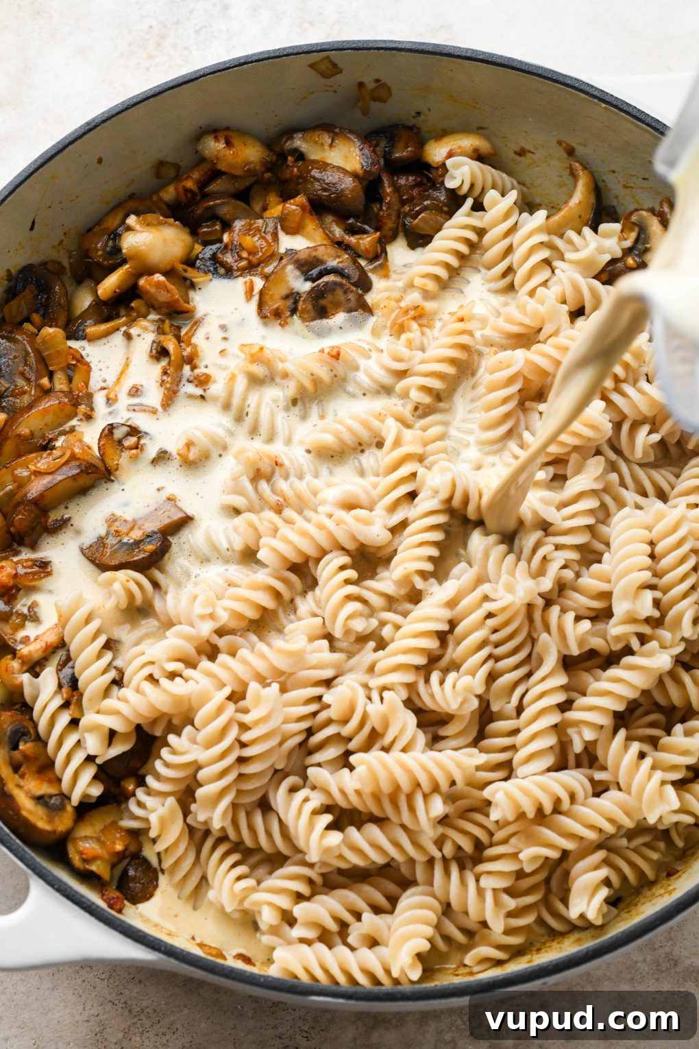 Cashew Cream Mushroom Stroganoff (Gluten-Free) 10 How to make creamy vegan mushroom stroganoff: Pouring cashew cream sauce into a large ceramic skillet with cooked mushrooms and pasta.