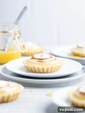Lemon meringue tartlet on a white plate with other tarts and a jar of lemon curd around it.