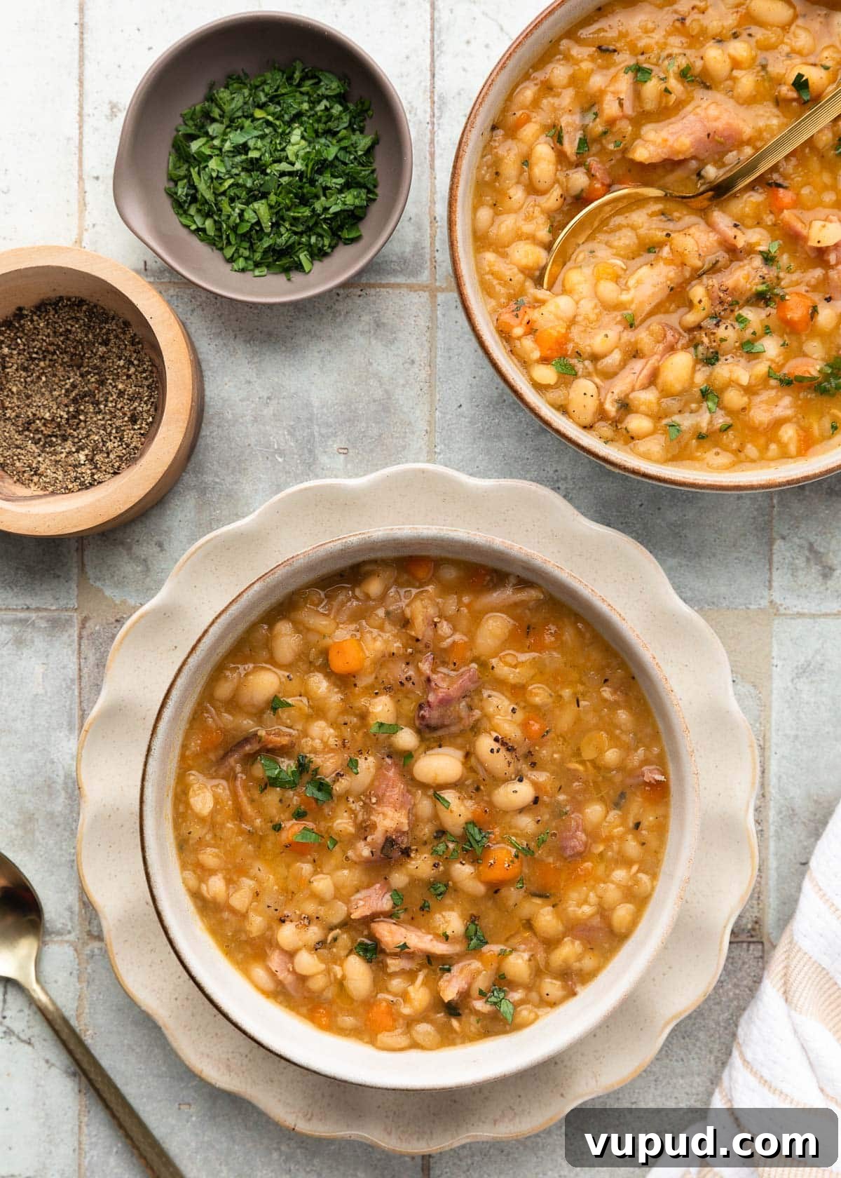 Classic Ham and Bean Soup Recipe for Comforting Weeknight Meals 13 overhead of two bowls of ham and bean soup on a gray tile surface next to smaller bowls of chopped parsley and ground black pepper