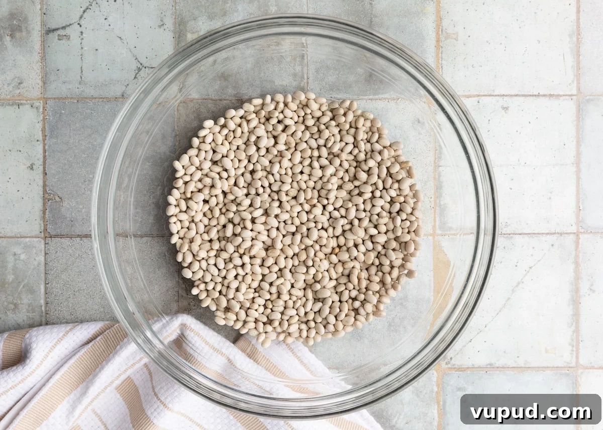 Classic Ham and Bean Soup Recipe for Comforting Weeknight Meals 4 dried navy beans in a glass bowl on a gray tile surface next to a white and ivory striped dish cloth