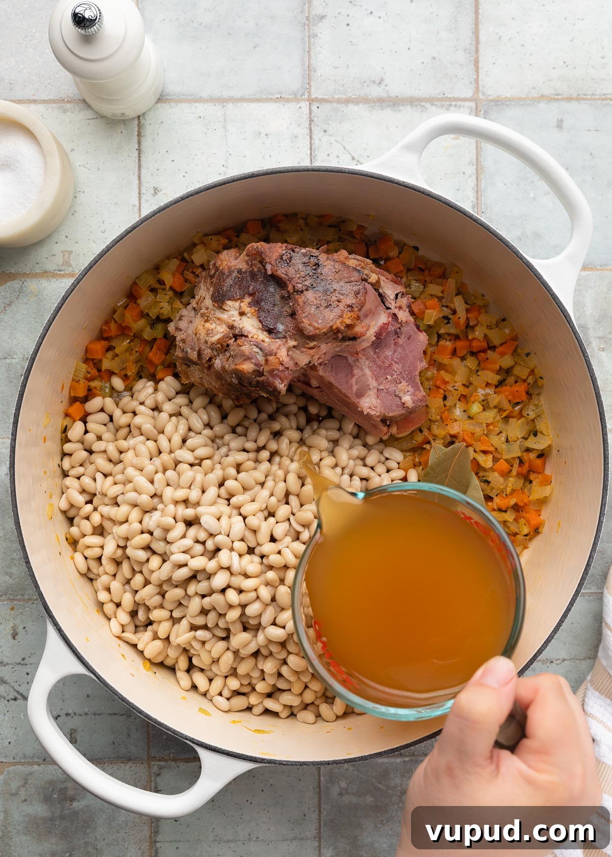 Classic Ham and Bean Soup Recipe for Comforting Weeknight Meals 8 chicken stock being poured from a liquid measuring cup into a white enameled cast iron Dutch oven with mirepoix, navy beans, and a ham bone