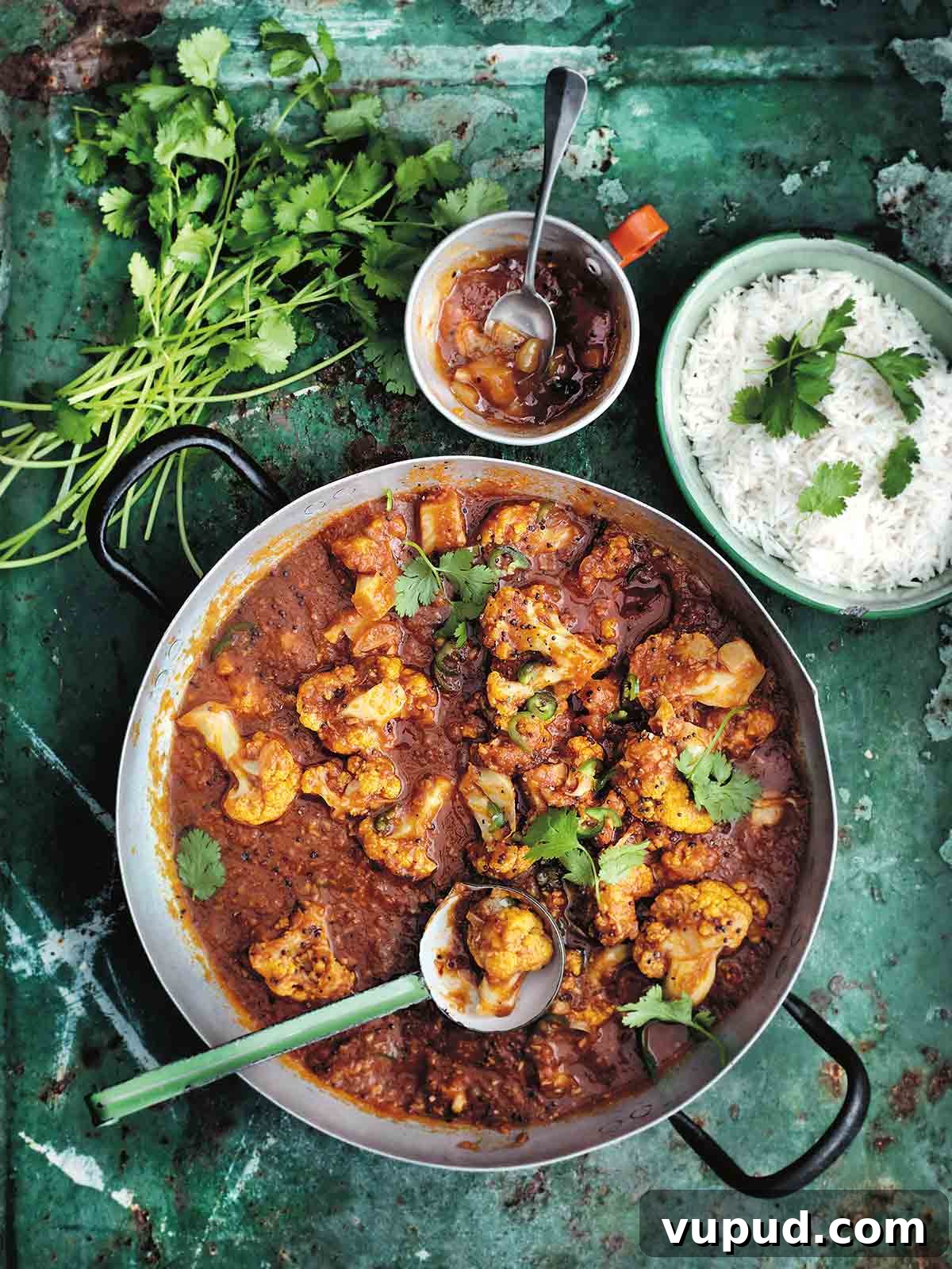 Cauliflower Tomato Curry Recipe: Flavorful Indian-Style Stew 2 A metal pot filled with cauliflower and tomato curry with a ladle resting inside, and a bowl of rice, a bowl of chutney and a pile of cilantro stems on the side.