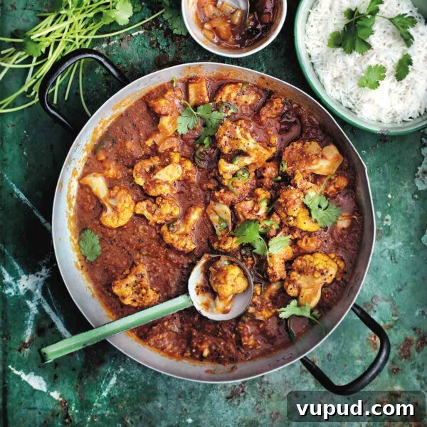 A metal pot filled with cauliflower and tomato curry with a ladle resting inside, and a bowl of rice, a bowl of chutney and a pile of cilantro stems on the side.