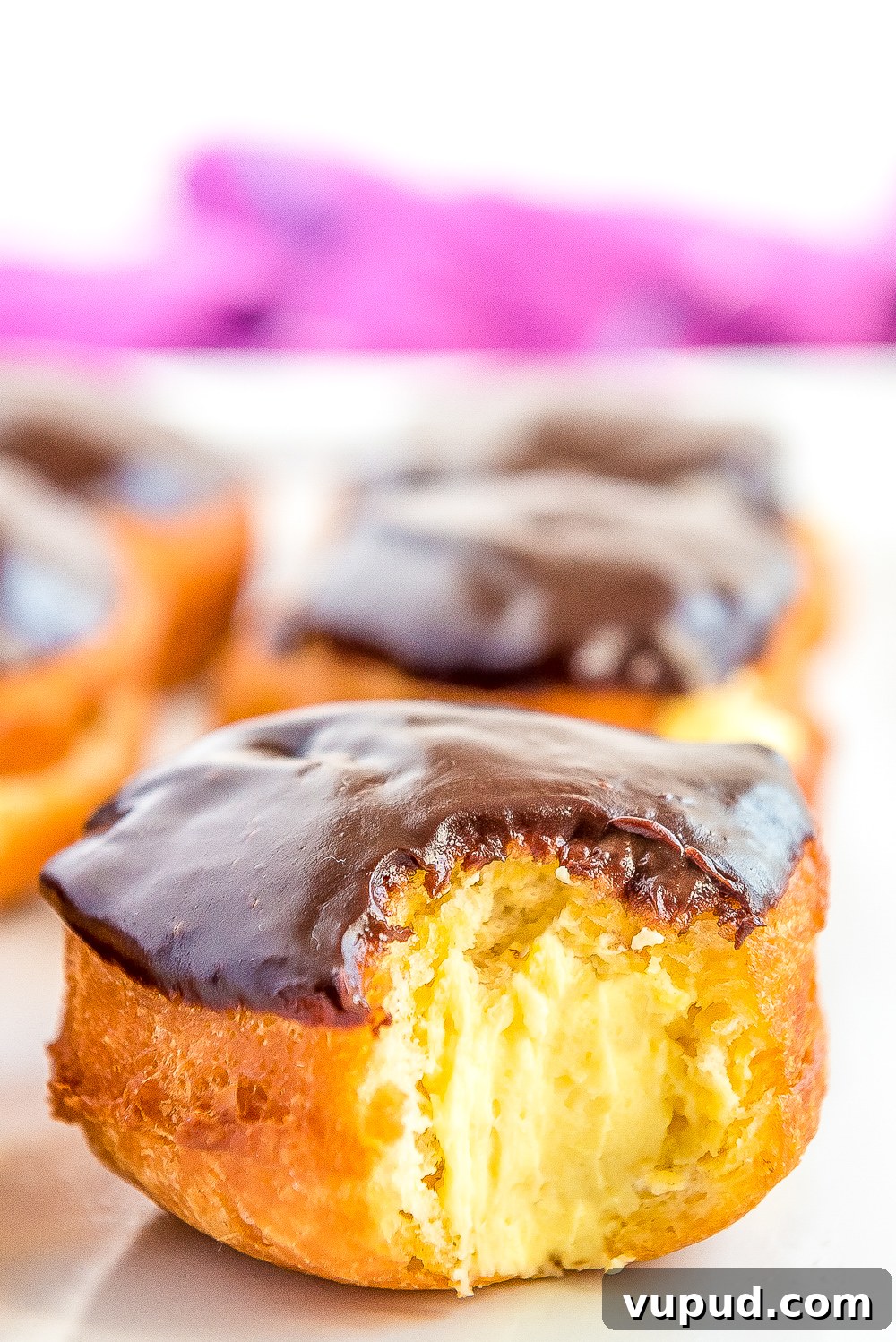 Boston Cream Donuts: Where to Find & How They're Made in Boston 3 Boston Cream Donut on white plate with a bite taken out of it showing the filling inside.