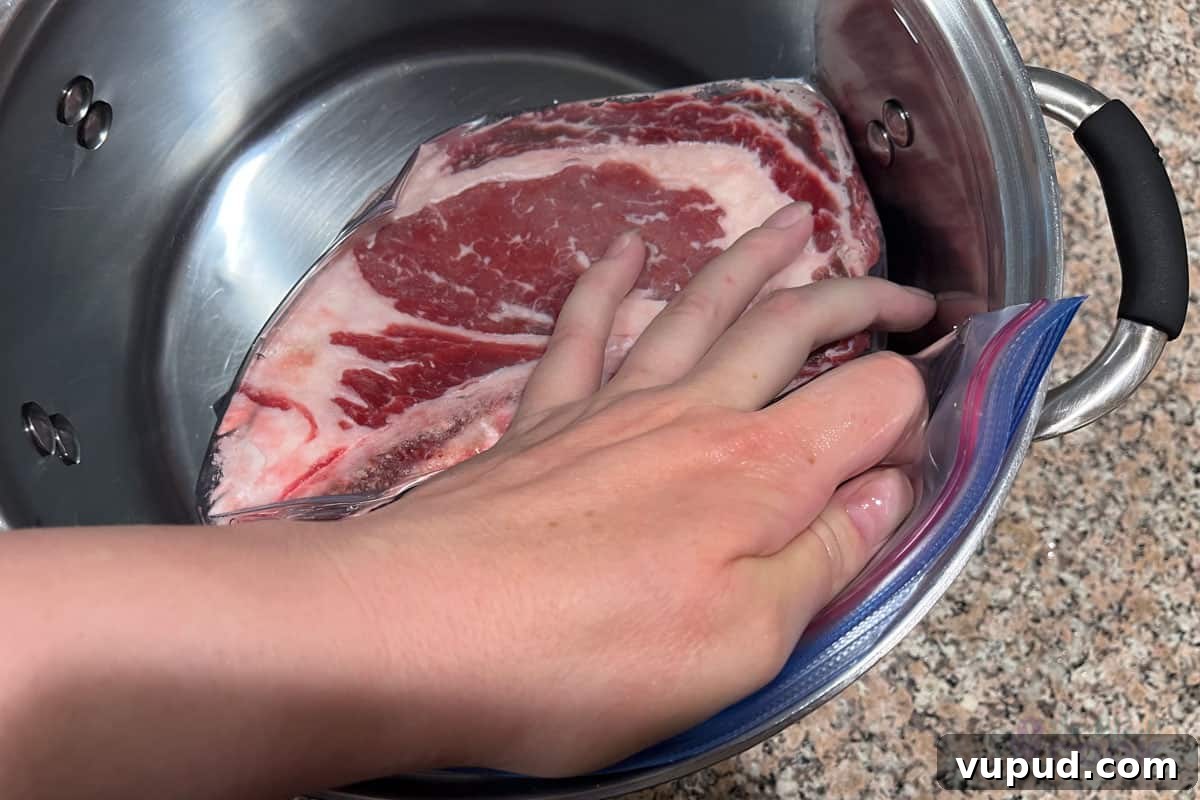 Perfectly Tender Sous Vide Prime Rib Roast Recipe 5 Rib roast in a gallon zip top bath with a hand holding it mostly underwater while pushing the air out the quarter of the seal that is still open.