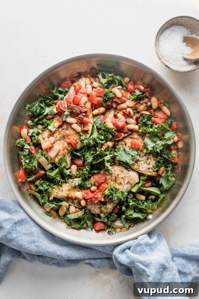 Tuscan Chicken with White Beans and Kale Recipe 2 Skillet full of a Tuscan-inspired chicken dinner with kale, white beans, and sun-dried tomatoes, with salt and pepper in the background for added seasoning.