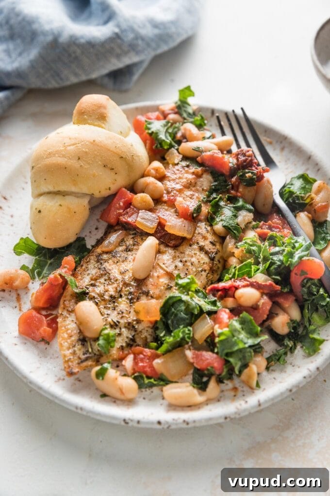 Tuscan Chicken with White Beans and Kale Recipe 7 Small plate with a portion of chicken with white beans, kale, and sun-dried tomatoes, served with a garlic knot.