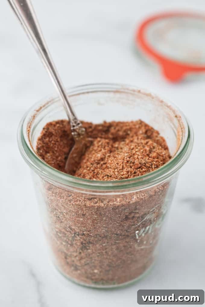 Homemade Cajun Seasoning Blend for Authentic Louisiana Flavor 2 Small glass mason jar filled with brownish red cajun spice seasoning with a silver spoon dug into the spices, mason jar is sitting on a marble table with mason jar lid and orange gasket sitting in background, blurry