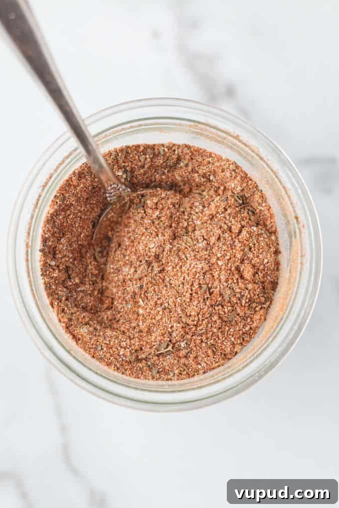 Homemade Cajun Seasoning Blend for Authentic Louisiana Flavor 6 Small glass mason jar filled with brownish red cajun spice seasoning with a silver spoon dug into the spices, mason jar is sitting on a marble table
