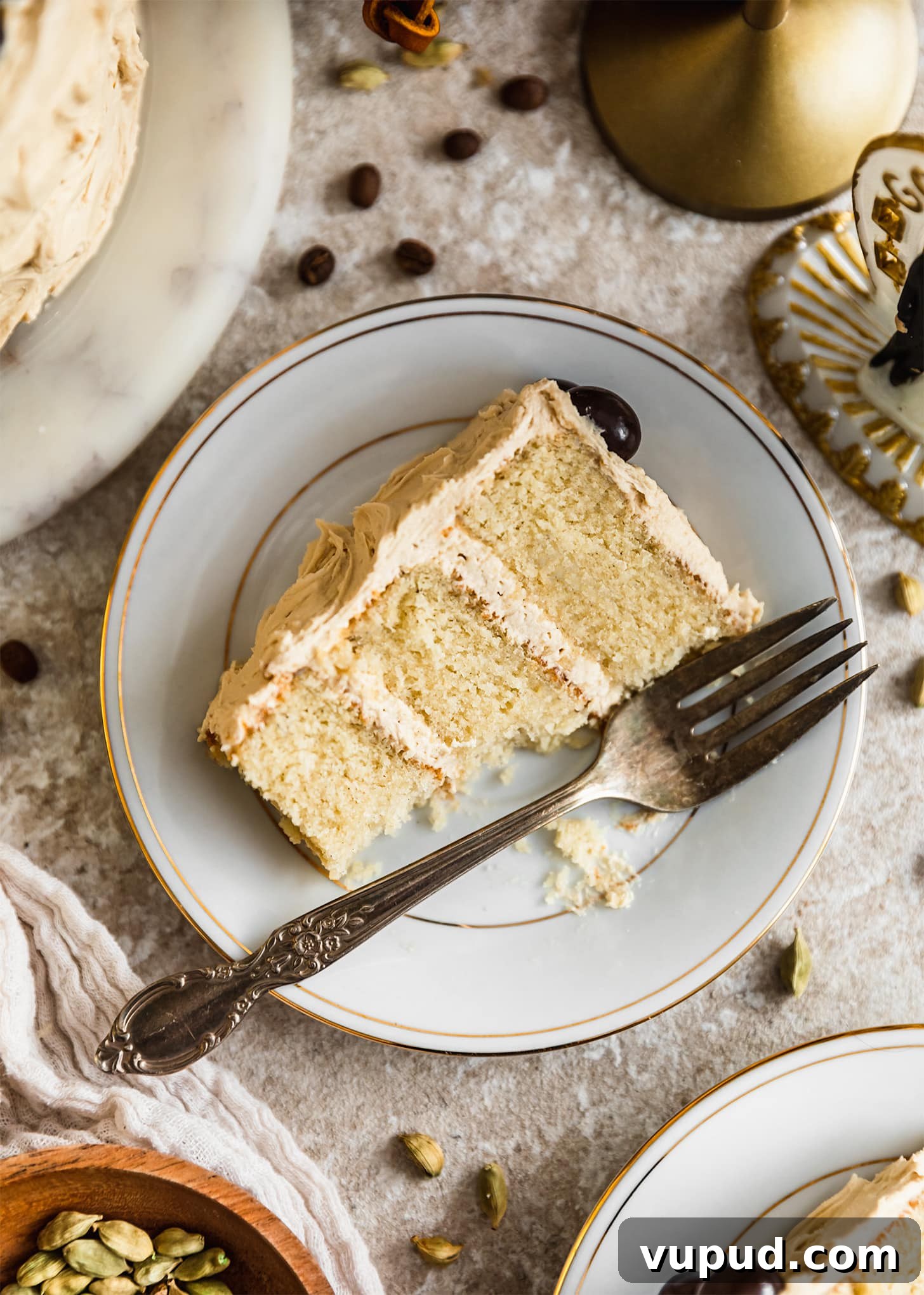 Cardamom Cake with Silky Coffee Buttercream 10 A white plate of cardamom cake with coffee frosting on a beige counter next to a white linen, wedding cake topper, coffee beans, and cardamom pods.