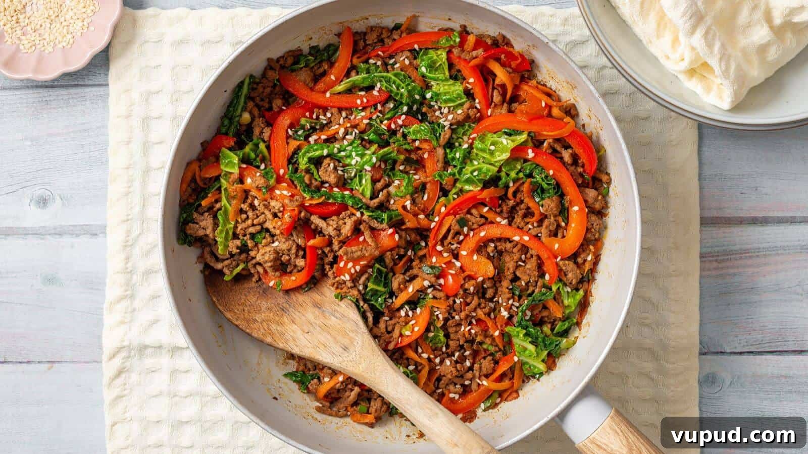 30 Flavorful Low-Carb Main Dishes That Feel Like Real Meals 30 A skillet containing a stir-fried mix of ground meat, sliced bell peppers, leafy greens, and white sesame seeds, with a wooden spoon resting on the side. Plates with flatbreads are in the background.