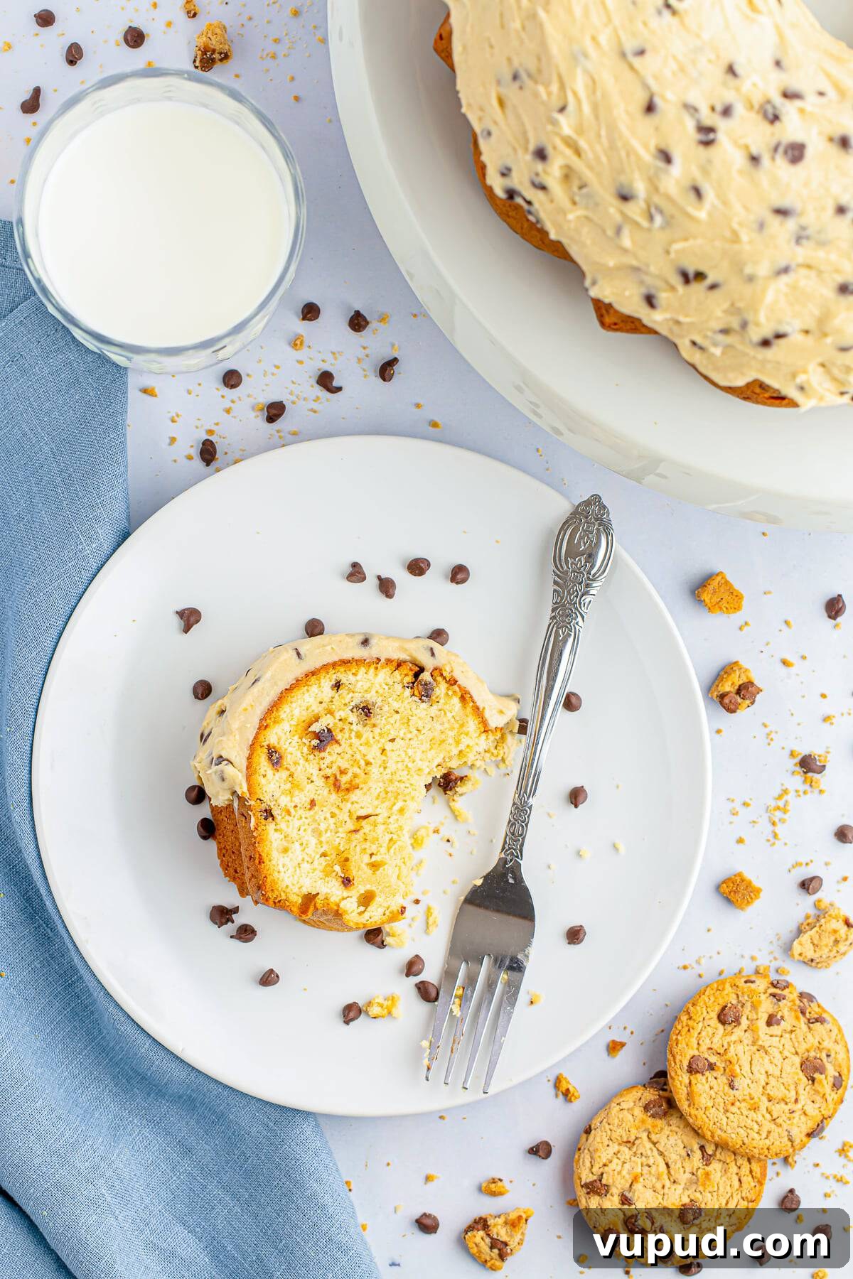 Chocolate Chip Bundt Cake with Cookie Dough Frosting Recipe 6 Overhead image of a slice of chocolate chip bundt cake on a white plate, the remainder of the cake on a white cake stand, glass of milk, blue linen and cookie crumbs