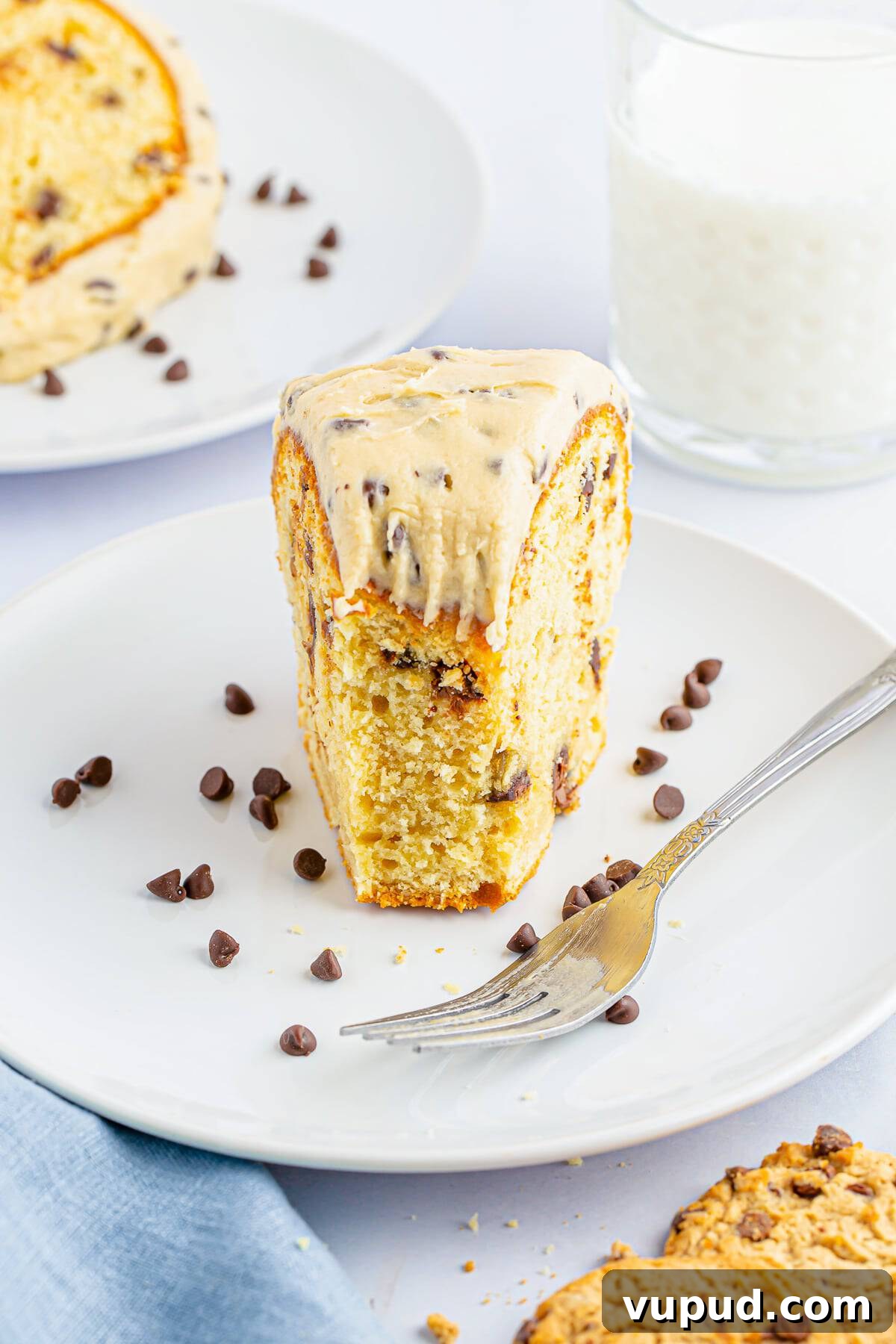 Chocolate Chip Bundt Cake with Cookie Dough Frosting Recipe 7 A bite taken out of a slice of chocolate chip bundt cake on a white plate with silver fork, glass of milk in the background, light blue linen, cookies, cookie crumbs, and chocolate chips scattered.