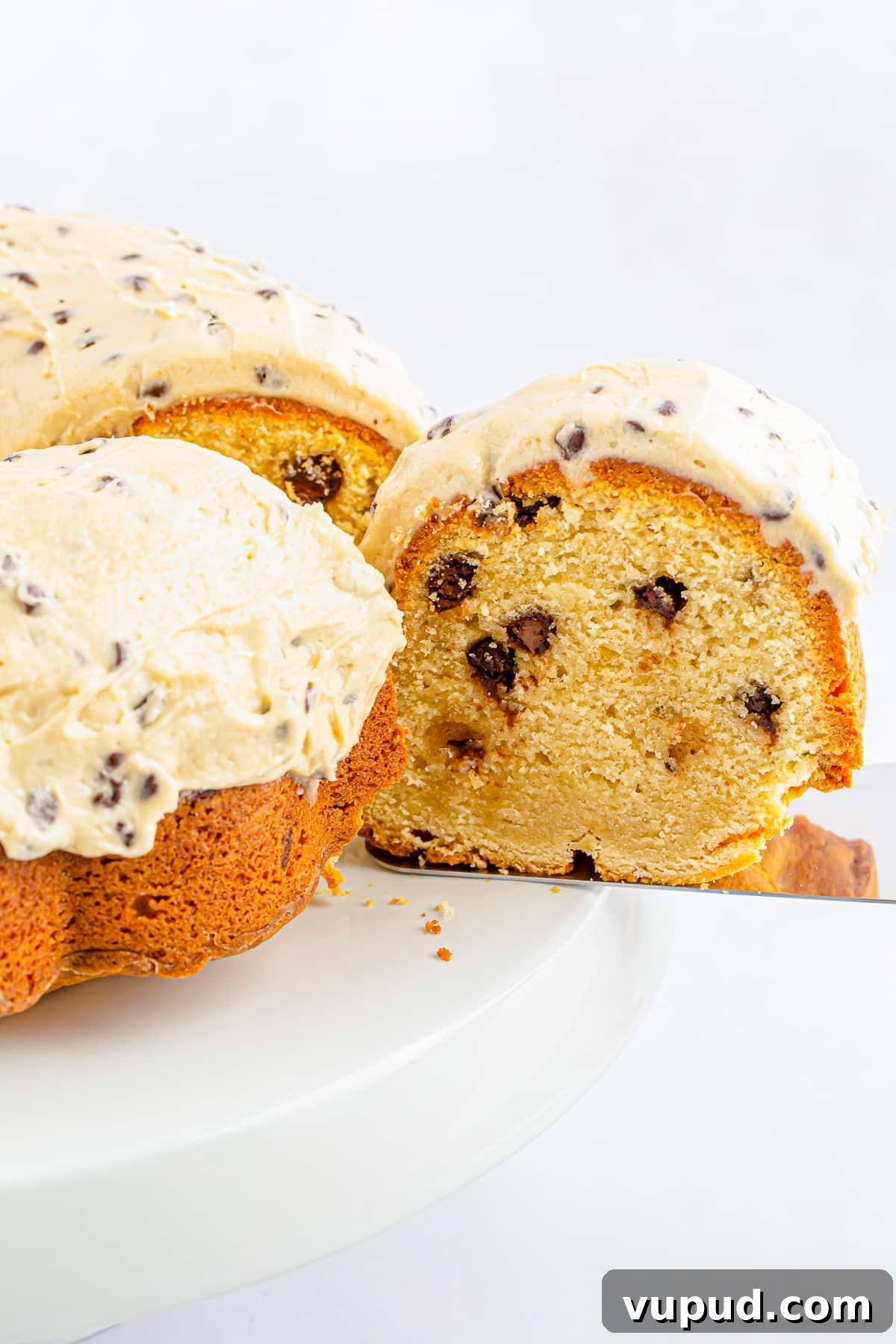 Chocolate Chip Bundt Cake with Cookie Dough Frosting Recipe 5 Upclose image of a slice of chocolate chip bundt cake coming out of the cake on a cake server, white cake stand