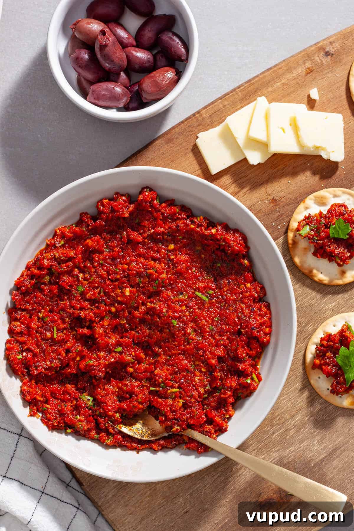 A bowl of sun-dried tomato spread with olives to the side and crackers on a cutting board.