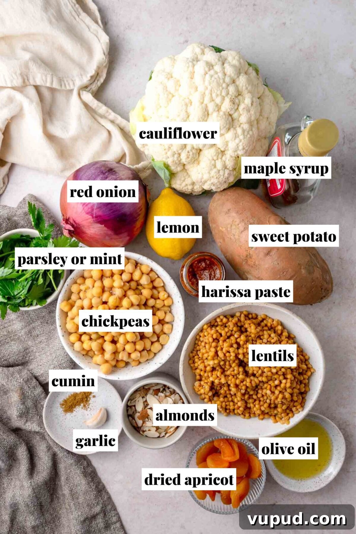Cauliflower, sweet potato, red onion, chickpeas, lentils, lemon, herbs, spices and oil arranged on a countertop.