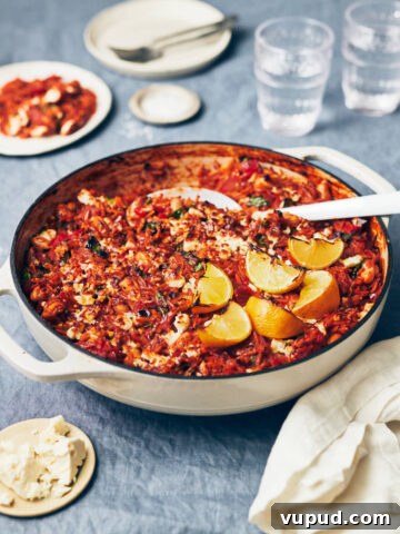 A white skillet filled with tomato lemon orzo on a table with smoked paprika, feta, and water glass in the background.